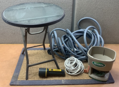 SMALL FLASHLIGHT (NO POWER) , METAL FRAME PATIO TABLE WITH GLASS TOP, A WHITE EXTENSION CORD, A GREY GARDEN HOSE WITH A SPRINKLER ATTACHMENT, SCOTTS WHIRL HAND HELD SPREADER W/ OUTDOOR MAT; PLEASE INSPECT