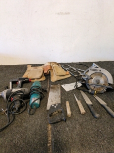 corded drill and another corded power tool, possibly a sander or grinder. handsaw and several knives or chisels with wooden handles. tool belt or pouch, and various other smaller tools like hacksaw blades and files.
