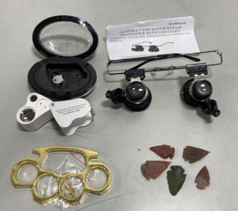 (1) Dual Magnification, Illuminated, Jewelers Loupe W/ Case, (1) Pair Watch Repair Magnifying Glasses W/ Light, (1) Brass Toned Paper Weight W/Belt Buckle Conversion Attachment, (1) Bag Of Assorted Carved Arrowheads